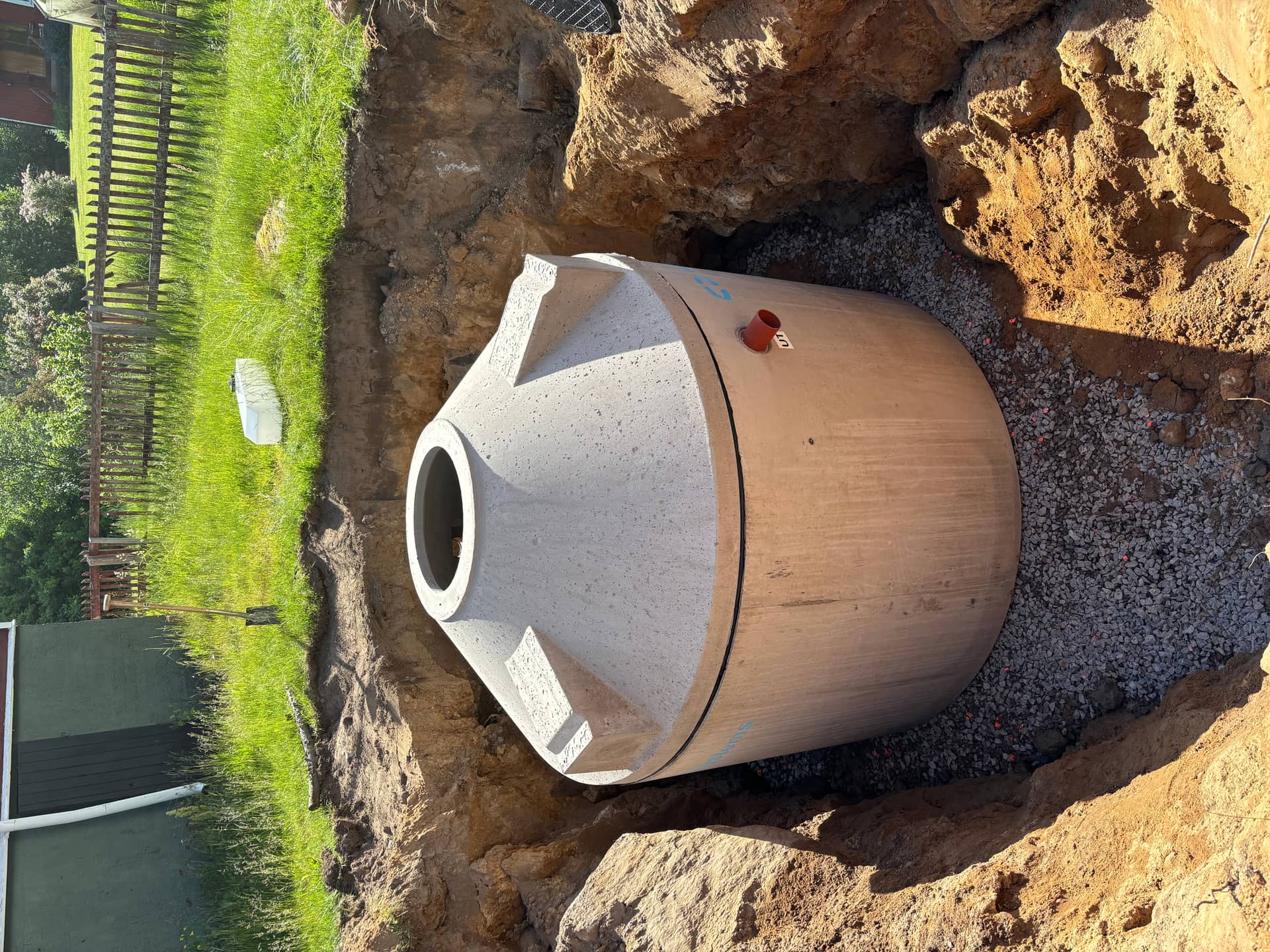 Betongtank med konisk topp och orange utlopp märkt UT i schakt på grusbädd