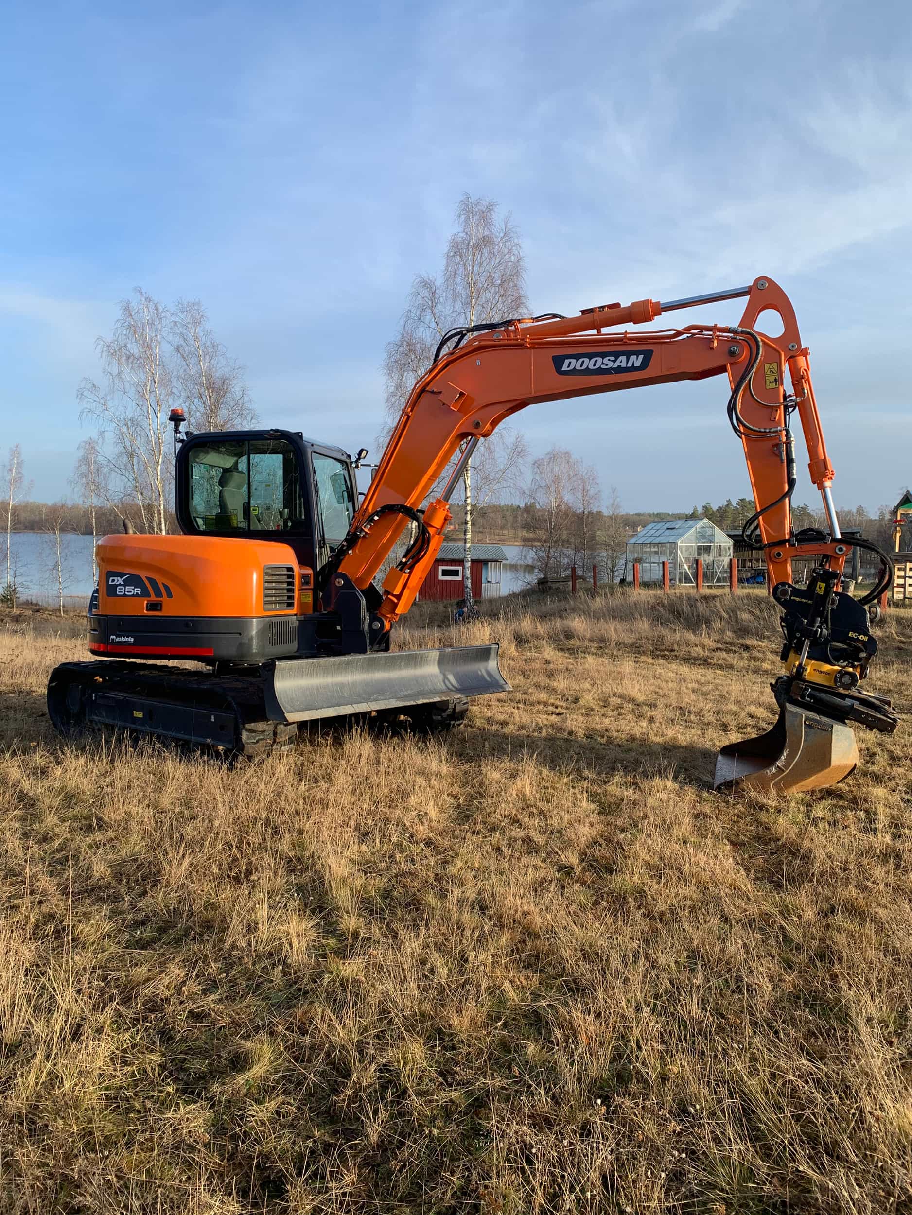 Orange Doosan DX85R-grävmaskin i profil på ett fält vid vatten, med tiltrotator och släntskopa