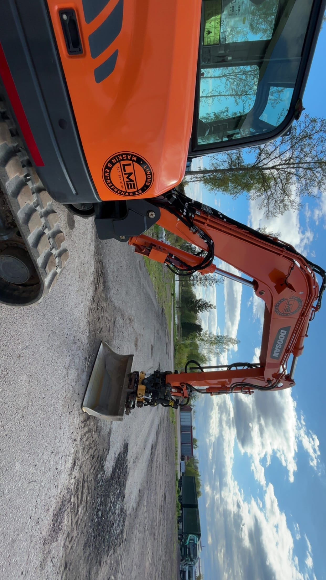 Doosan-grävmaskin i närbild med LME-logotyp och släntskopa mot vatten och träd