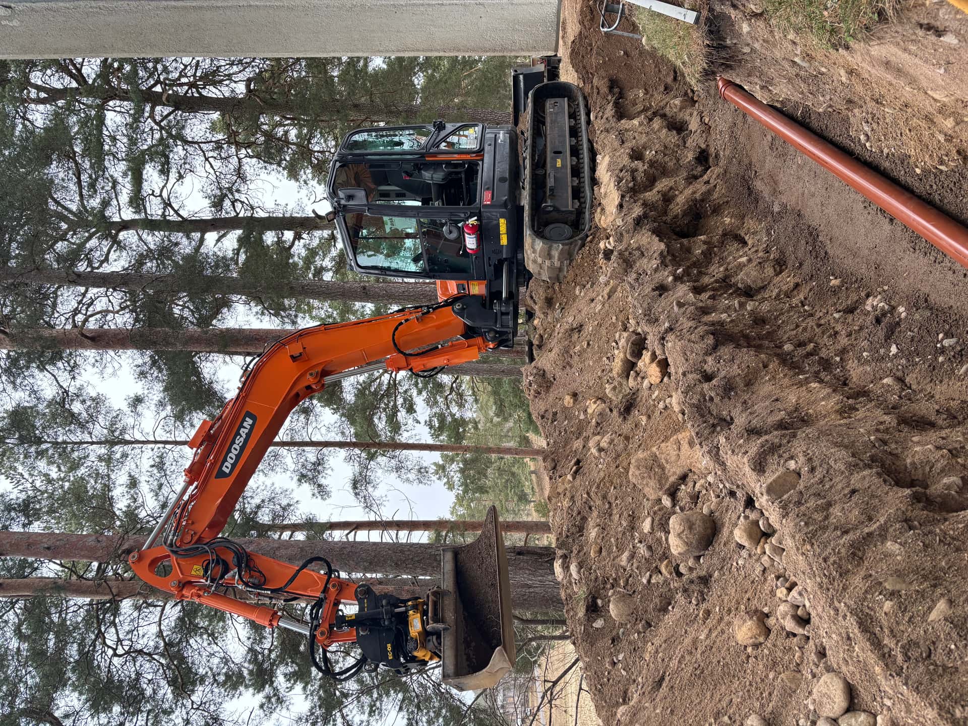 Doosan-grävmaskin vid djupt schakt med brun avloppsledning i sandbädd