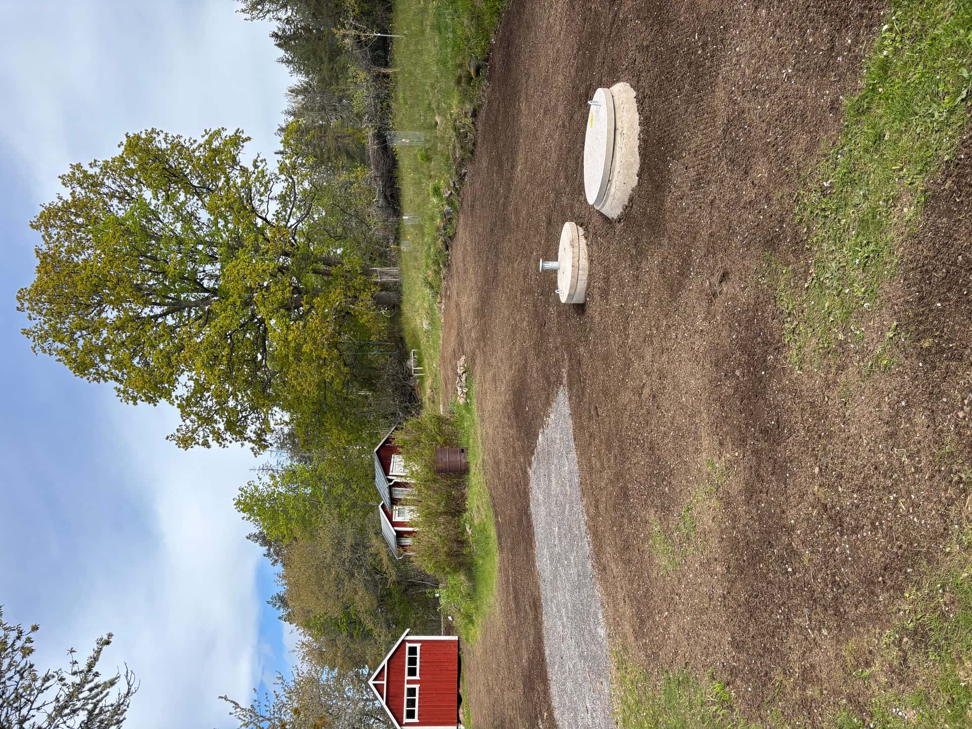 Utlagd matjord med två betongbrunnslock framför röda bodar och ett stort lövträd
